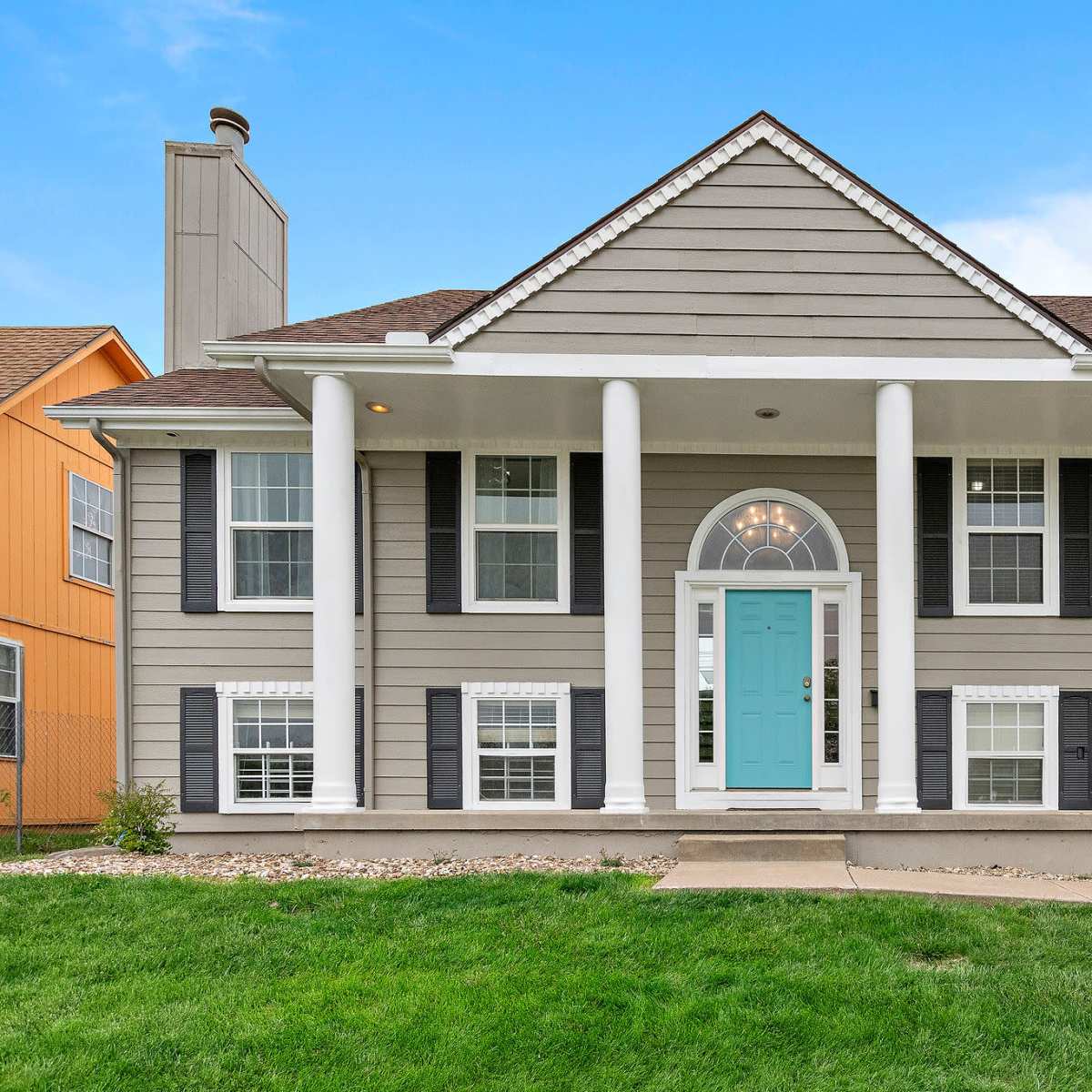 A two-story gray house with white columns and a turquoise front door, surrounded by a well-maintained lawn.