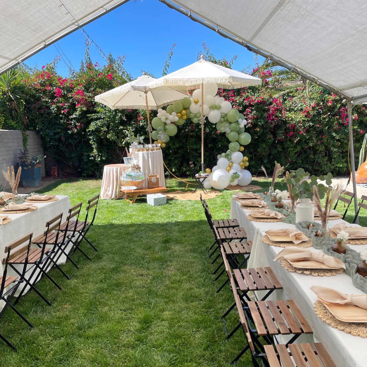 A tented outdoor setting features long tables with beige tablecloths, folding chairs, and decorative umbrellas, surrounded by greenery and floral arrangements.