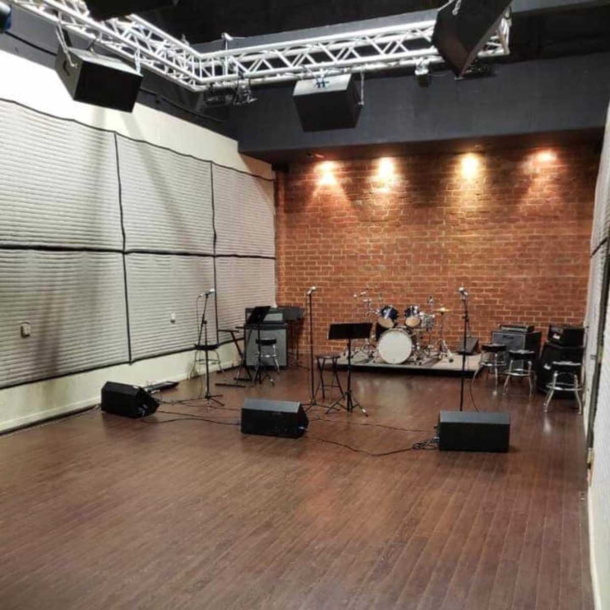 A music rehearsal studio with a wooden floor, brick walls, and various instruments set up, including a drum kit and microphones.
