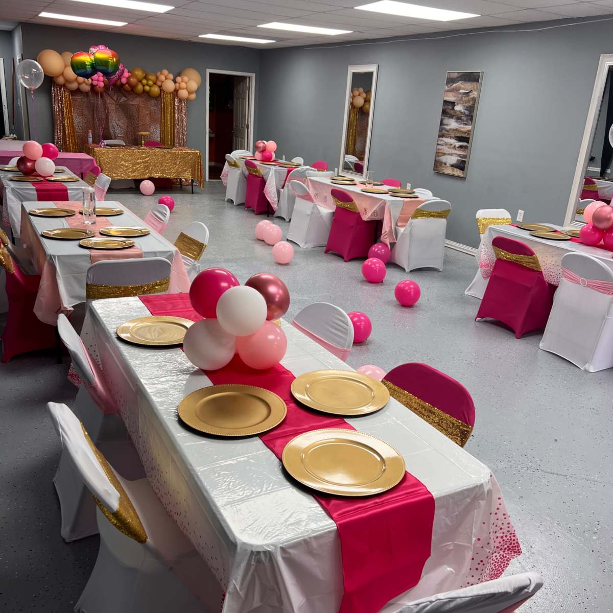 A banquet room arranged with several tables covered in white and pink tablecloths, decorated with gold plates and colorful balloons.