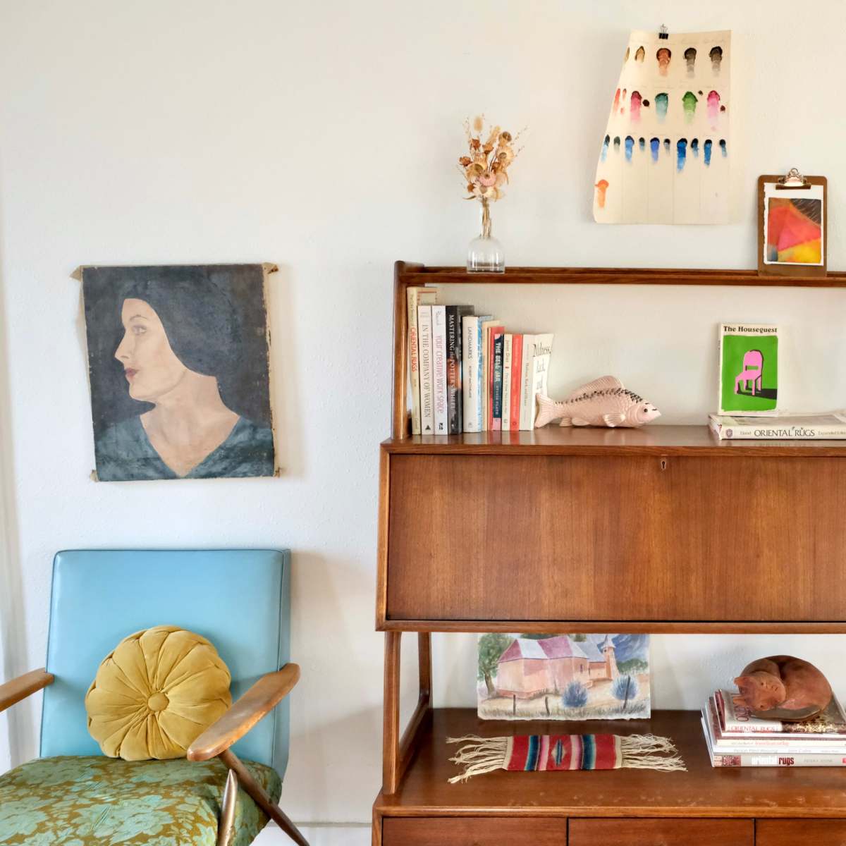A wooden bookshelf filled with books, decorative items, and framed artwork, positioned beside a chair in a well-lit room.