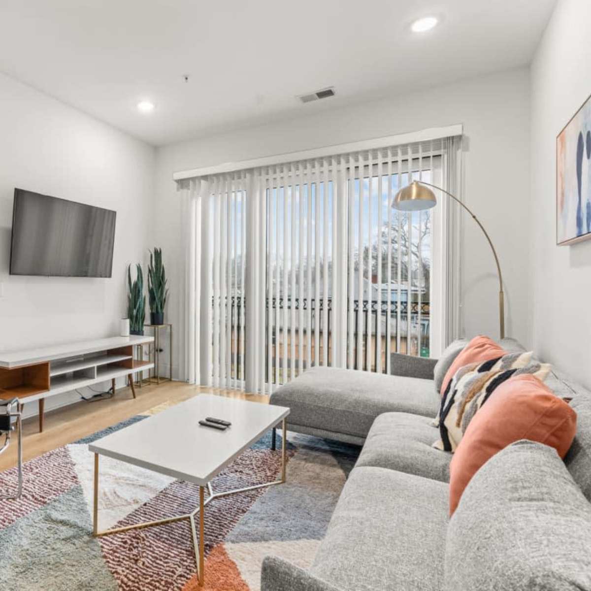 A modern living room with a sectional sofa, a coffee table, a flat-screen TV on the wall, and a sliding glass door leading to a balcony.