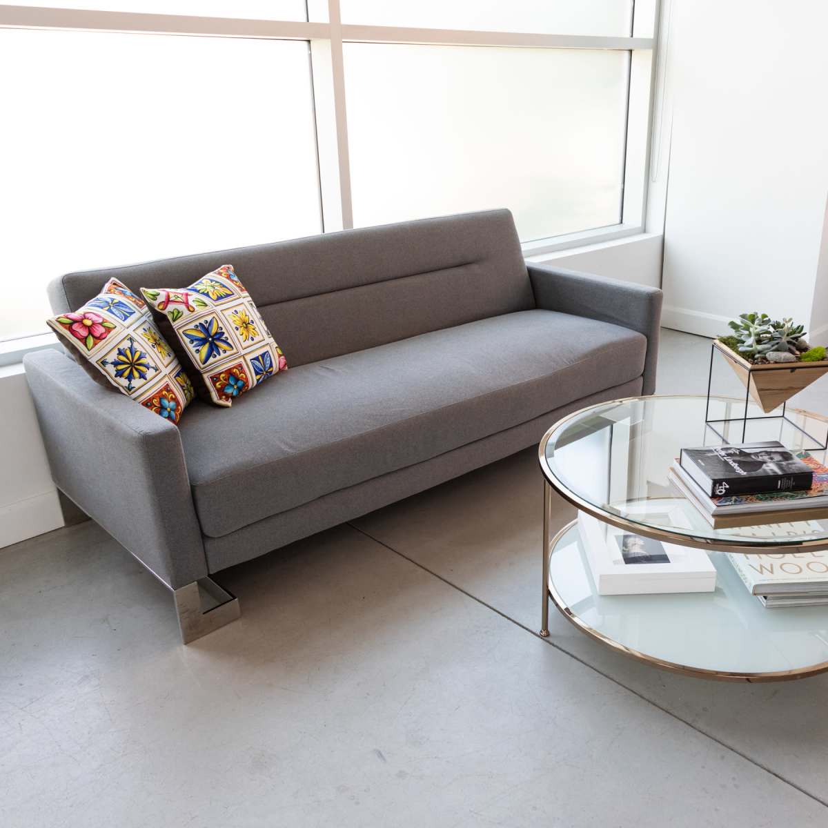 A gray sofa with decorative pillows is positioned next to a round glass coffee table holding books and a small plant in a modern interior space.
