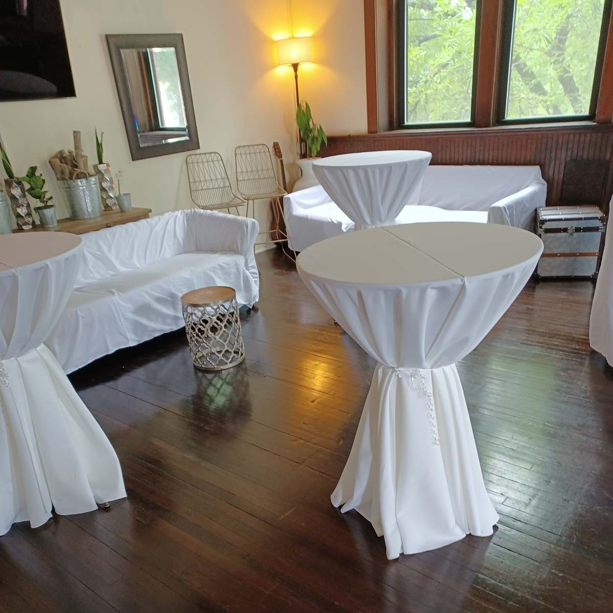 A room with wooden floors featuring several white-covered tables and a seating area with a couch and a decorative stool.
