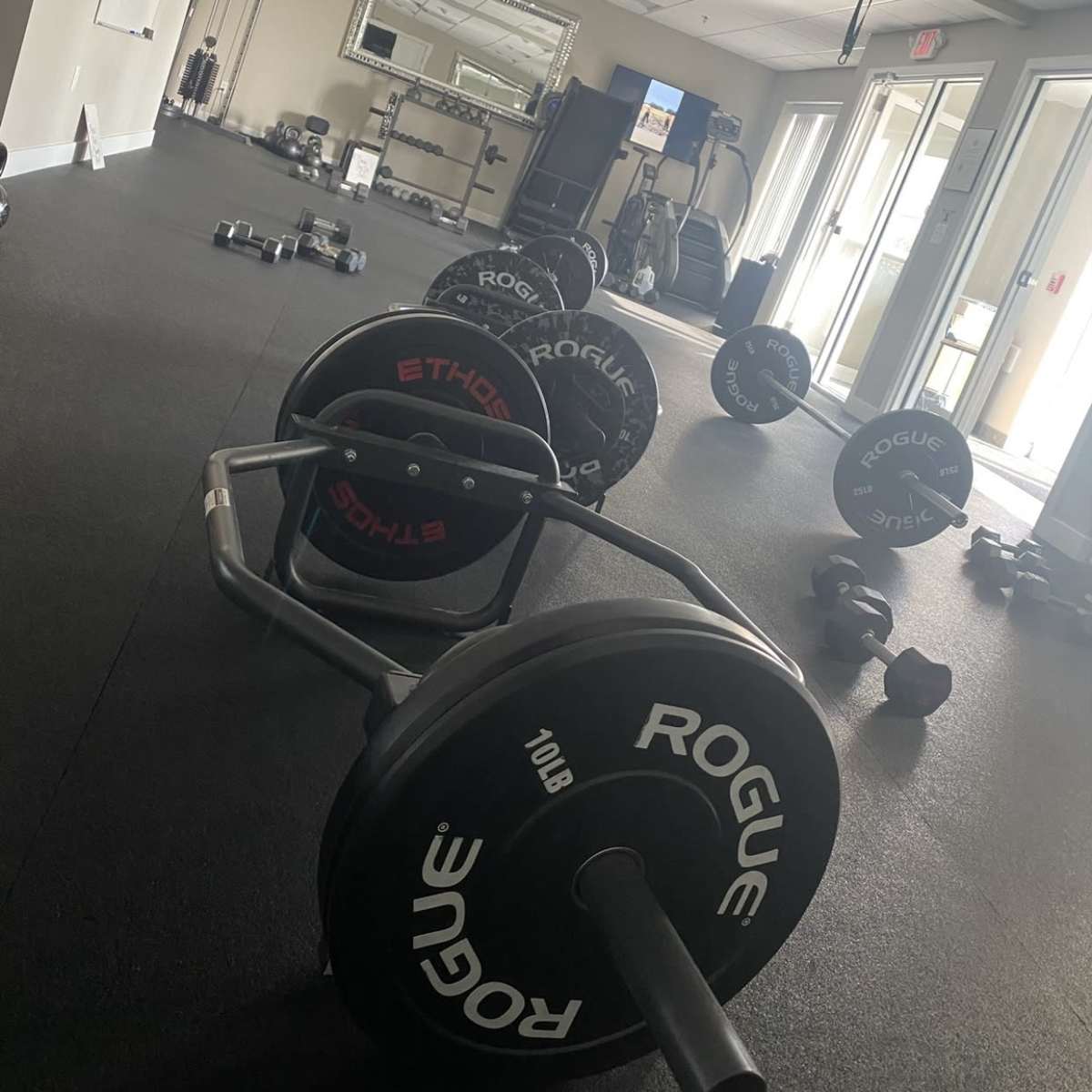 A fitness room with various weights, including barbells and dumbbells, arranged on a rubber floor, with workout equipment positioned in the background.