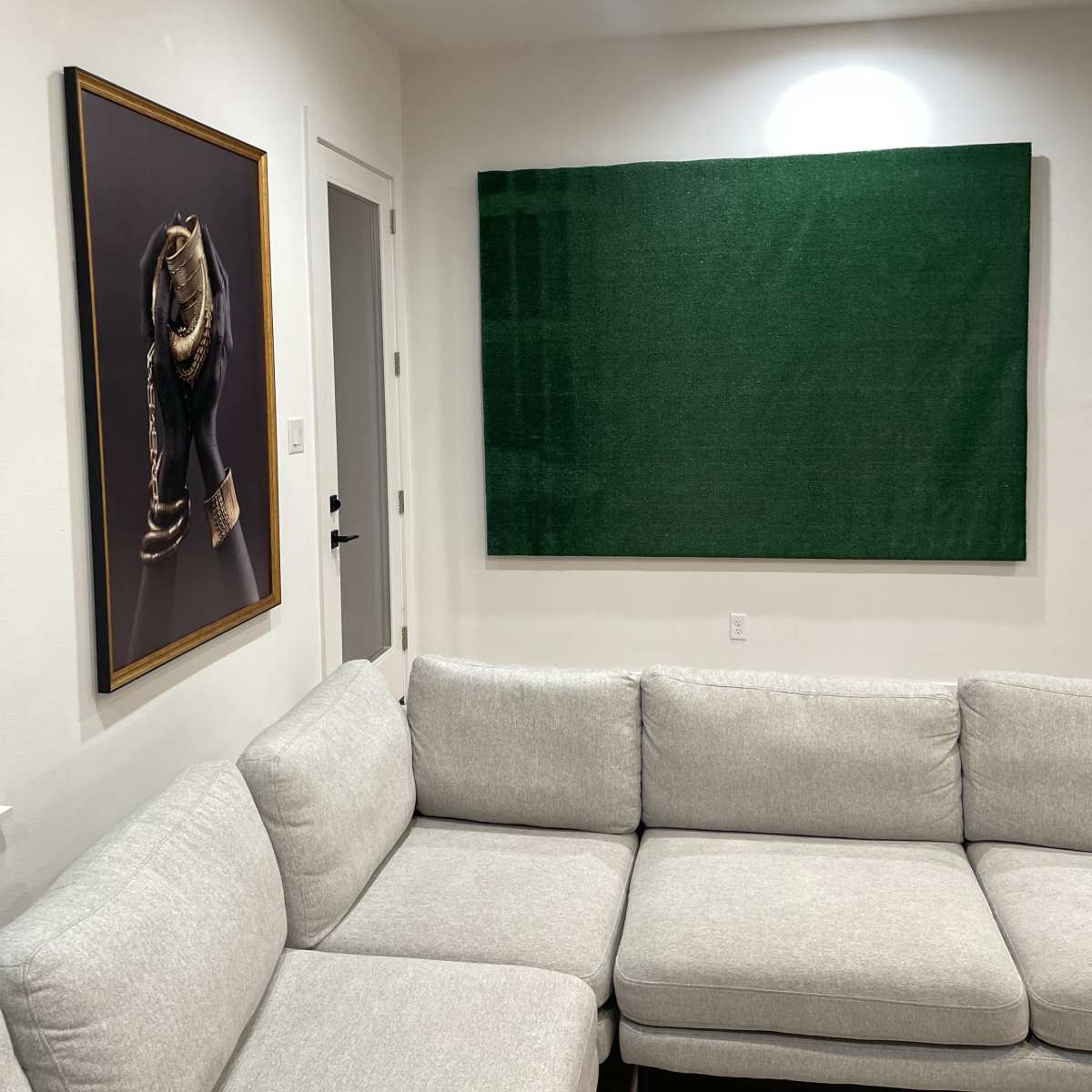 A cozy room with a sectional gray couch, a framed artwork of a baseball glove on the left wall, and a large green panel on the right wall.