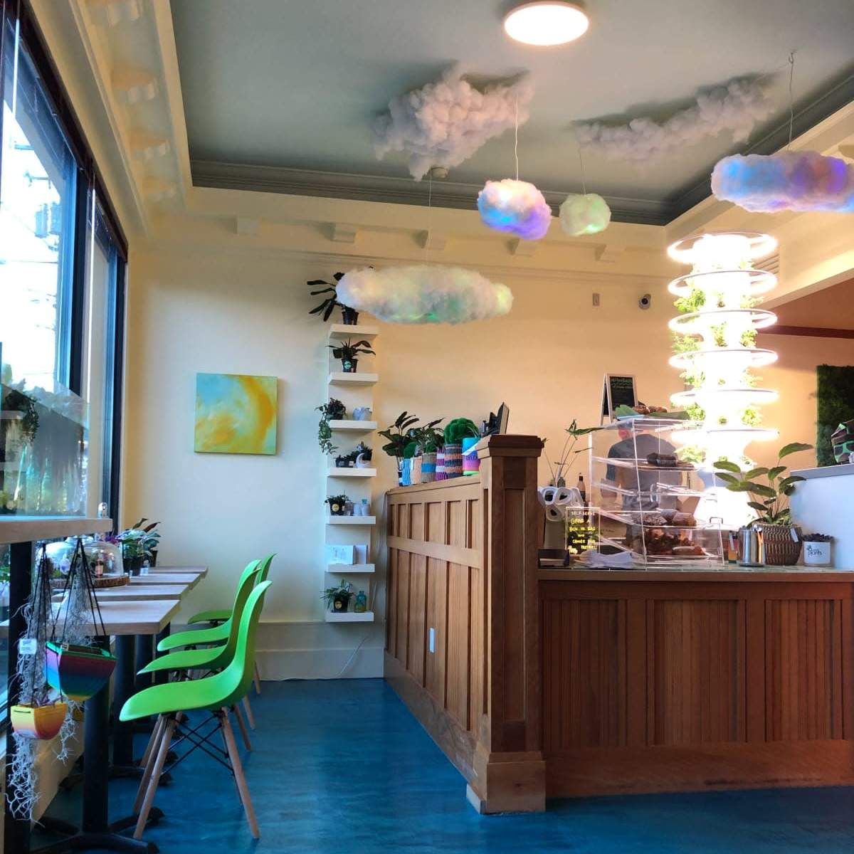 A brightly lit café interior with green chairs, plants on shelves, and a wooden counter decorated with hanging lights and cloud-shaped décor.