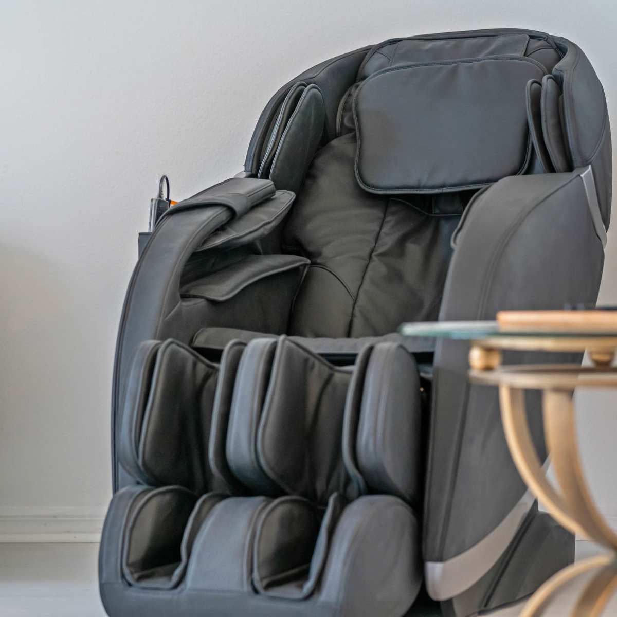 A black massage chair is positioned next to a small glass-topped side table in a light-colored room.