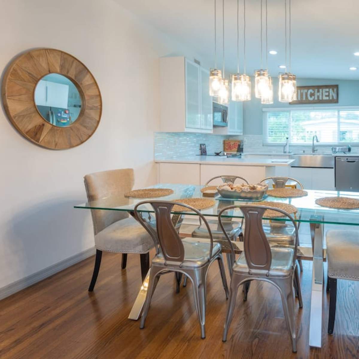 A modern dining area features a glass table with six chairs, a round mirror on the wall, and a kitchen visible in the background.