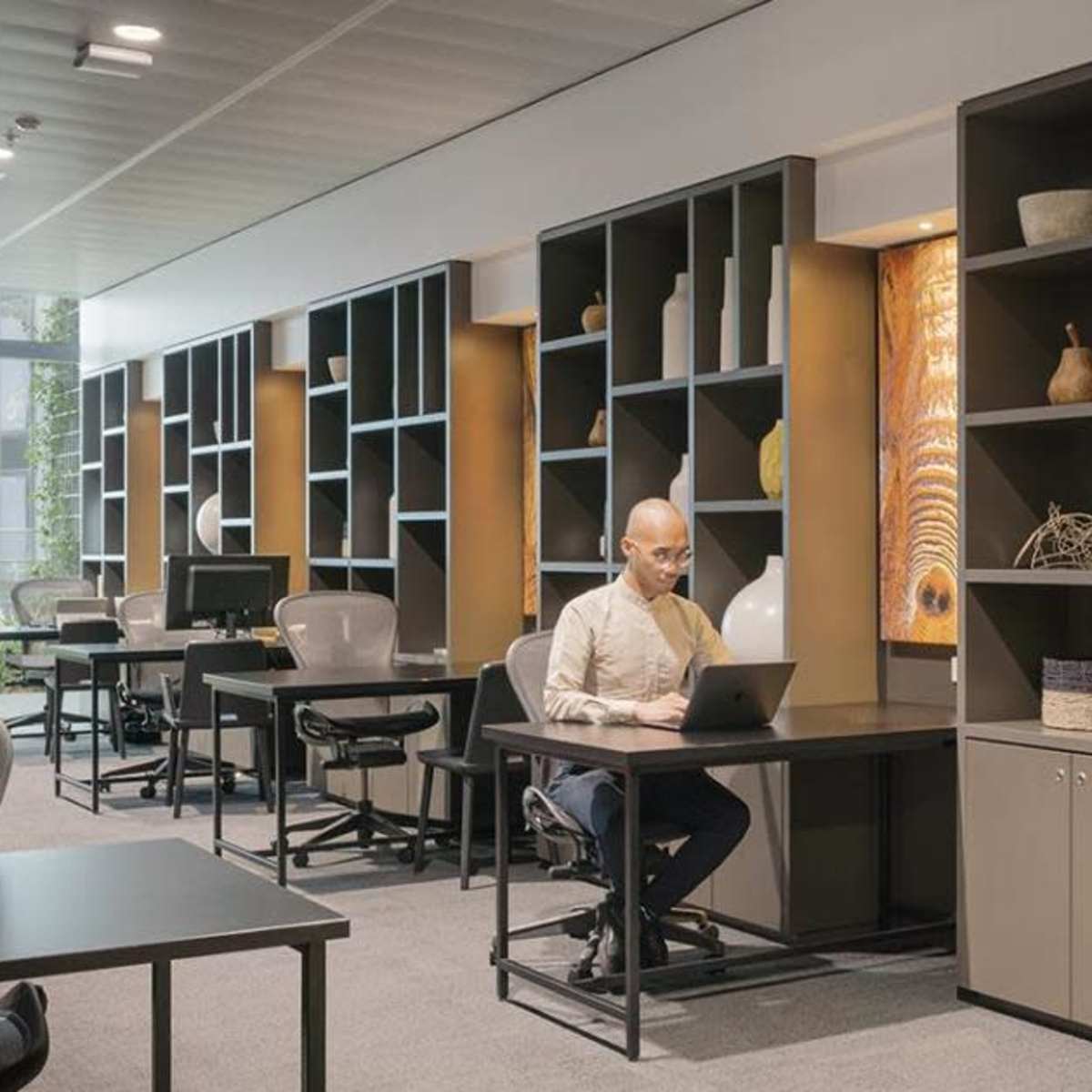 A workspace features a man working on a laptop at a desk, surrounded by shelves displaying various decorative items.
