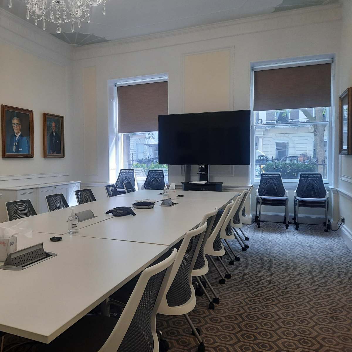 A conference room with a long white table, black chairs, a large screen on one wall, and framed portraits on the adjacent walls.