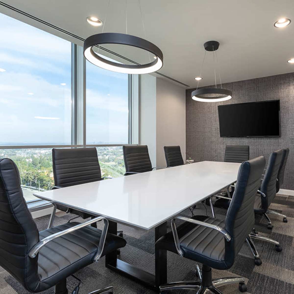 A modern conference room features a long white table surrounded by black leather chairs, with large windows offering a view and a wall-mounted TV.