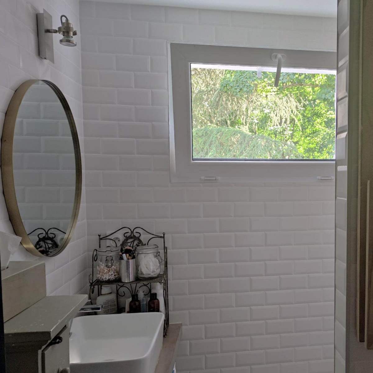 A bathroom with white tiled walls, a circular mirror, a sink, and a window providing natural light.