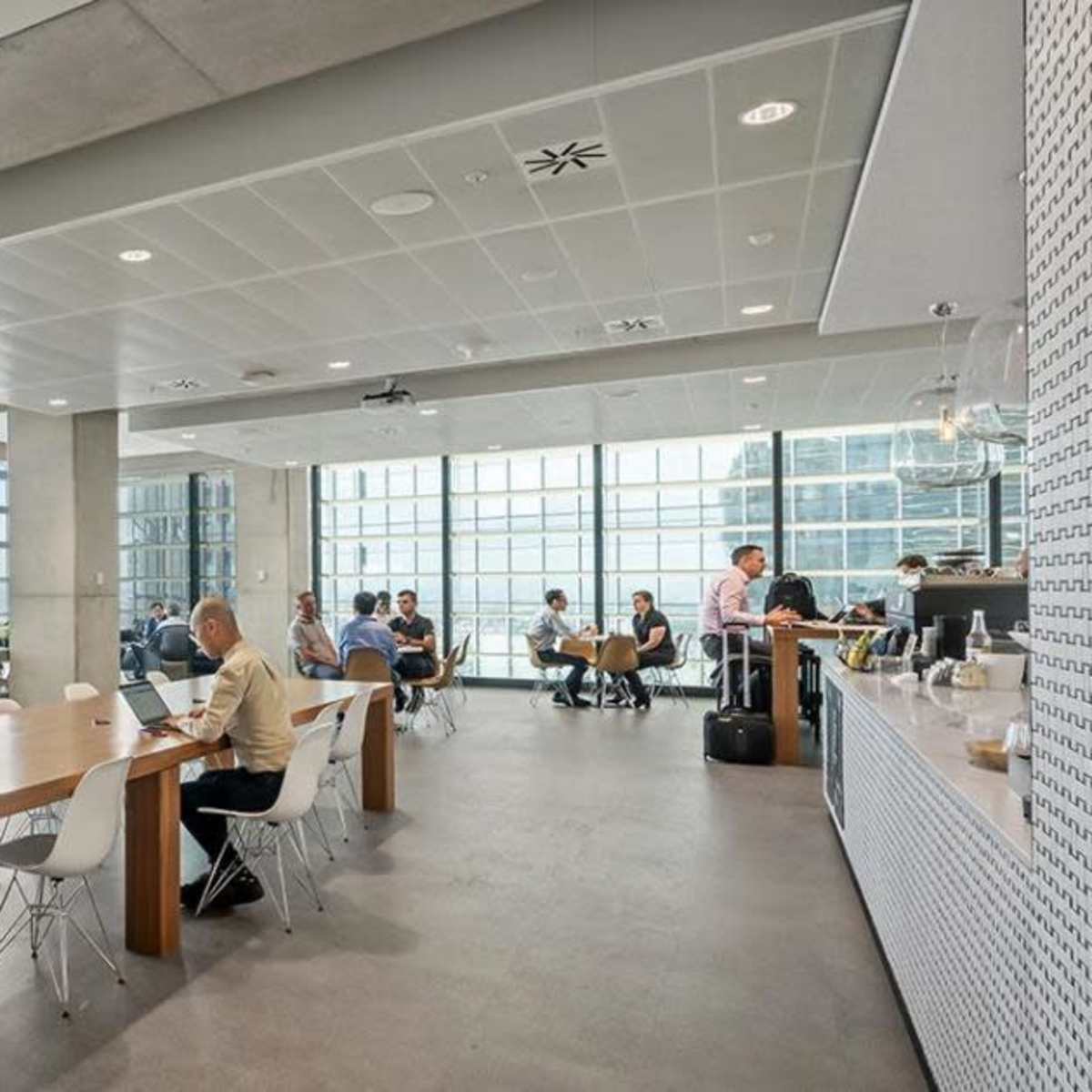 A modern office space with large windows, a long communal table, and several people engaged in work or conversation.