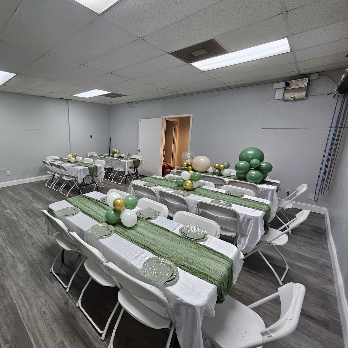A spacious event room is arranged with long tables covered in white cloth and green table runners, surrounded by white folding chairs and decorated with balloon clusters in various colors.