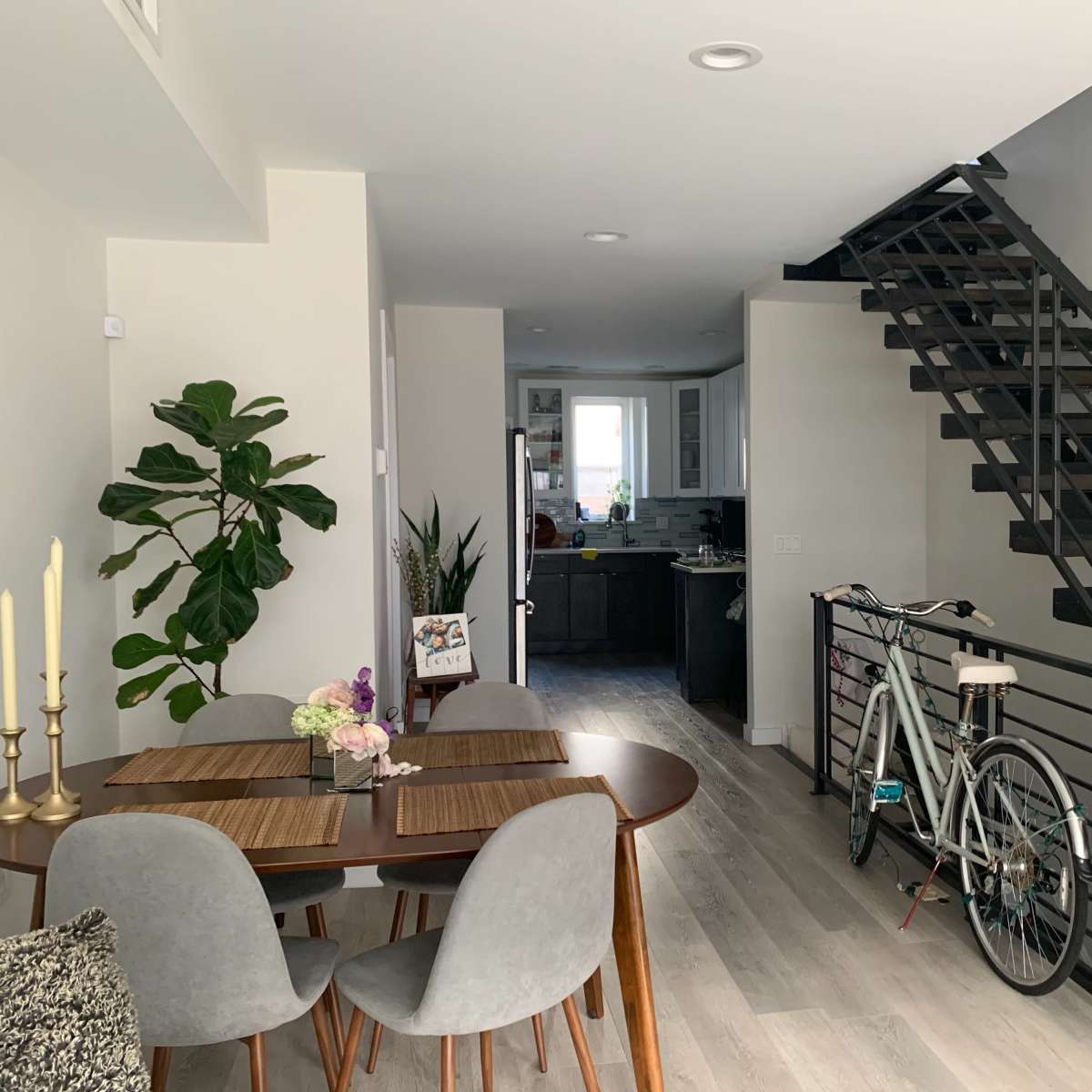 A dining area with a round table and chairs is positioned next to a staircase, while a bicycle is parked nearby and a doorway leads to a kitchen.