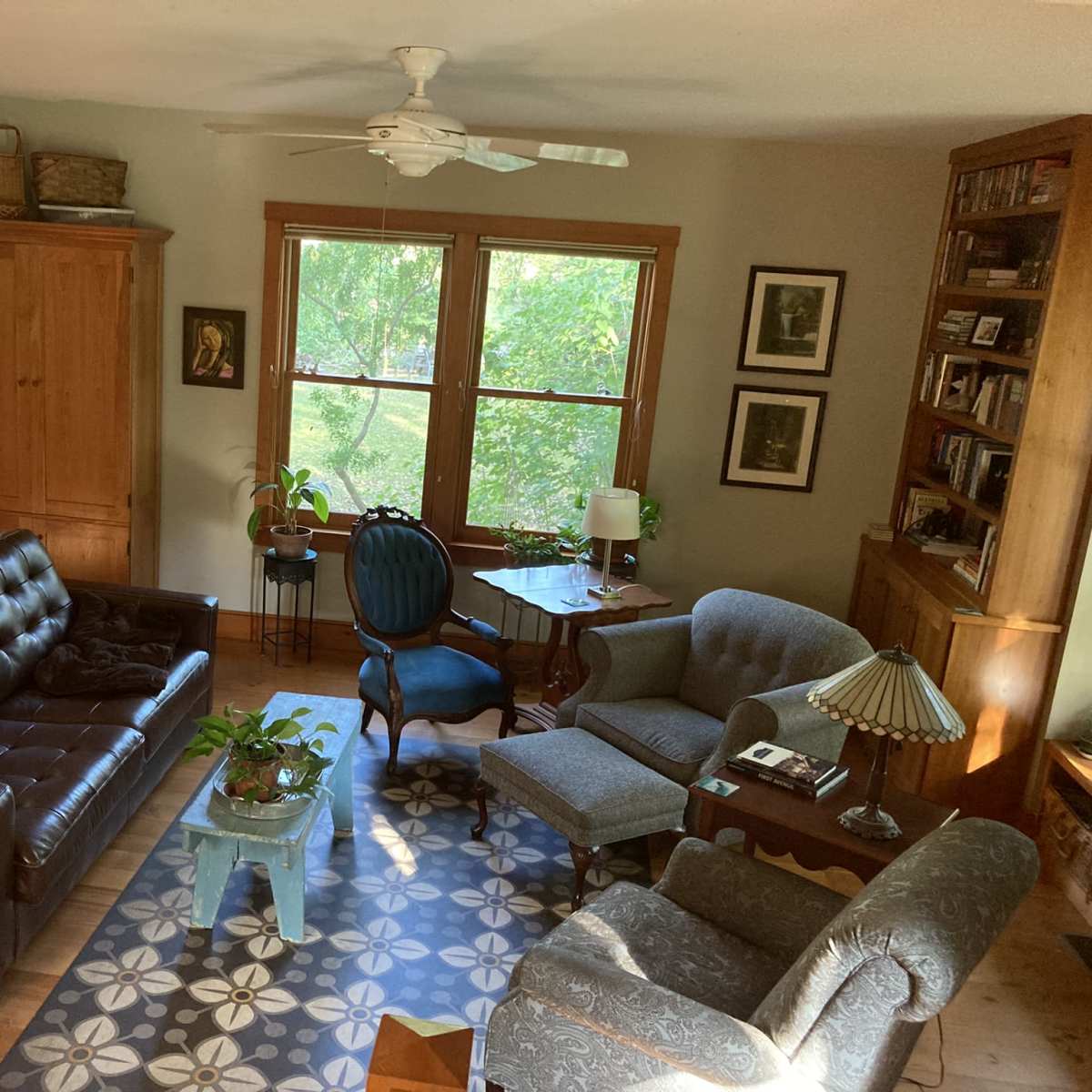A cozy living room with a brown leather sofa, a blue armchair, and a wooden bookshelf, illuminated by natural light from large windows.