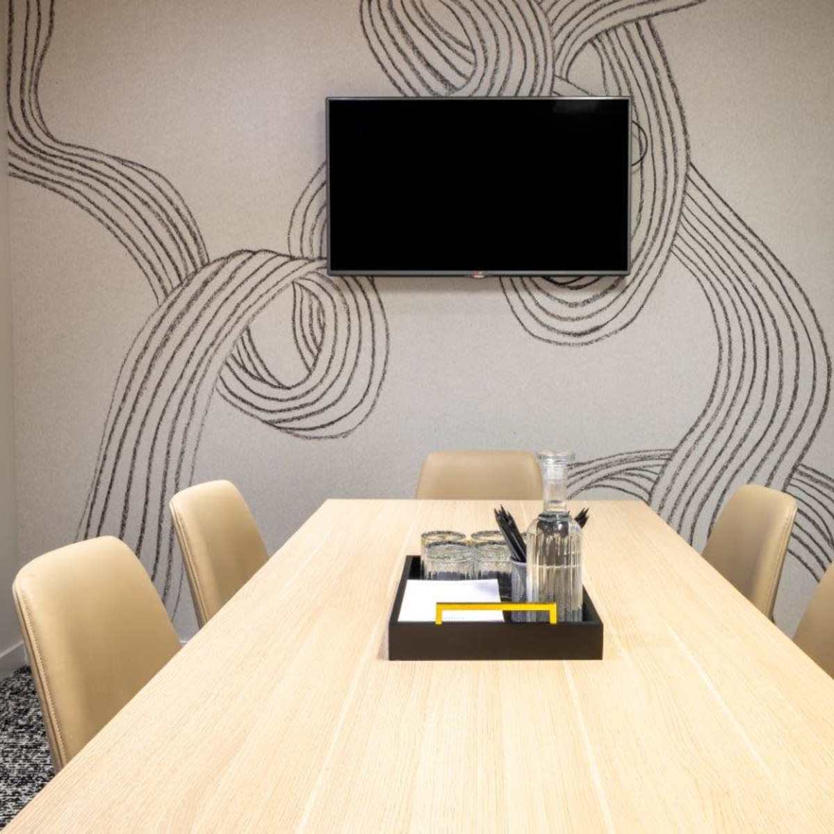 A conference room features a long wooden table surrounded by beige chairs, with a television mounted on a patterned wall and a coat rack in the corner.