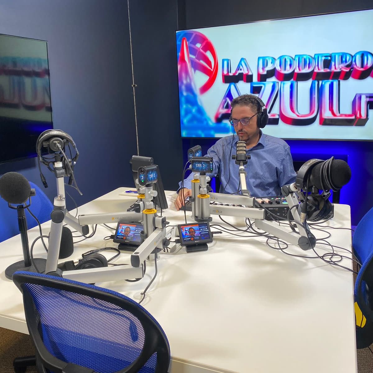 A radio host sits at a table equipped with microphones and audio equipment, surrounded by a blue-themed studio with a logo on the wall.