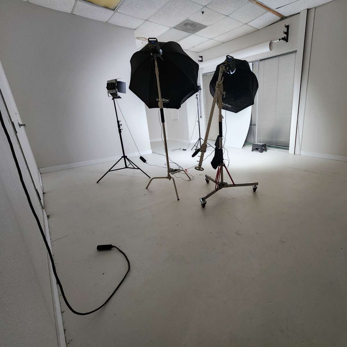 A sparsely furnished studio space with two softbox light stands and a tripod, set against white walls and a large window.