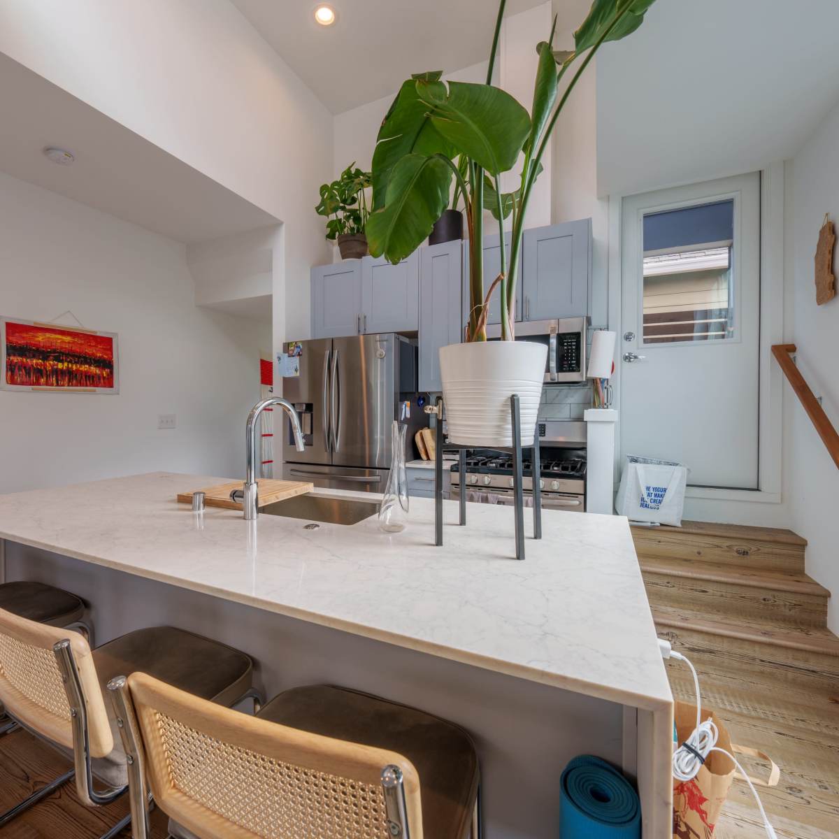 A modern kitchen features a large island with a marble countertop, bar stools, and light cabinetry, along with a potted plant beside a staircase and a window.