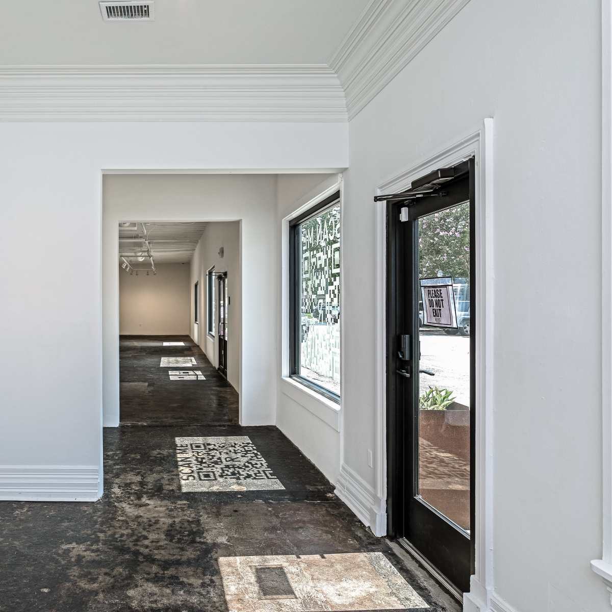 A long, narrow hallway with white walls, two doorways, and large windows along one side, revealing a view of the exterior.