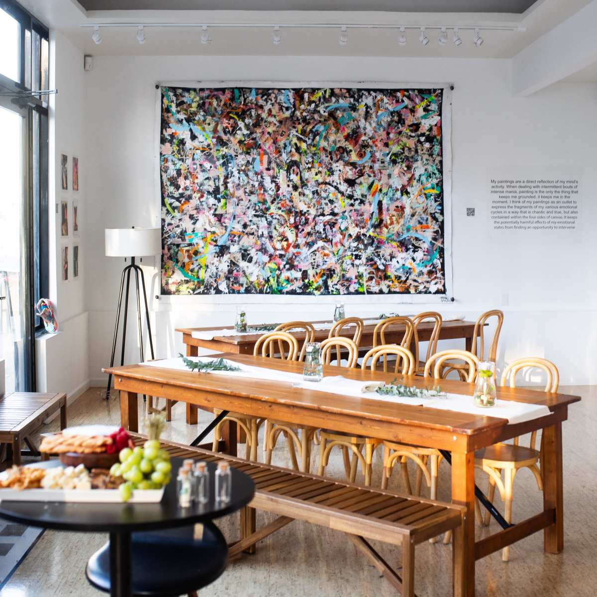 A spacious dining area features long wooden tables set with small vases, a large colorful abstract painting on the wall, and a side bar holding food and drinks.
