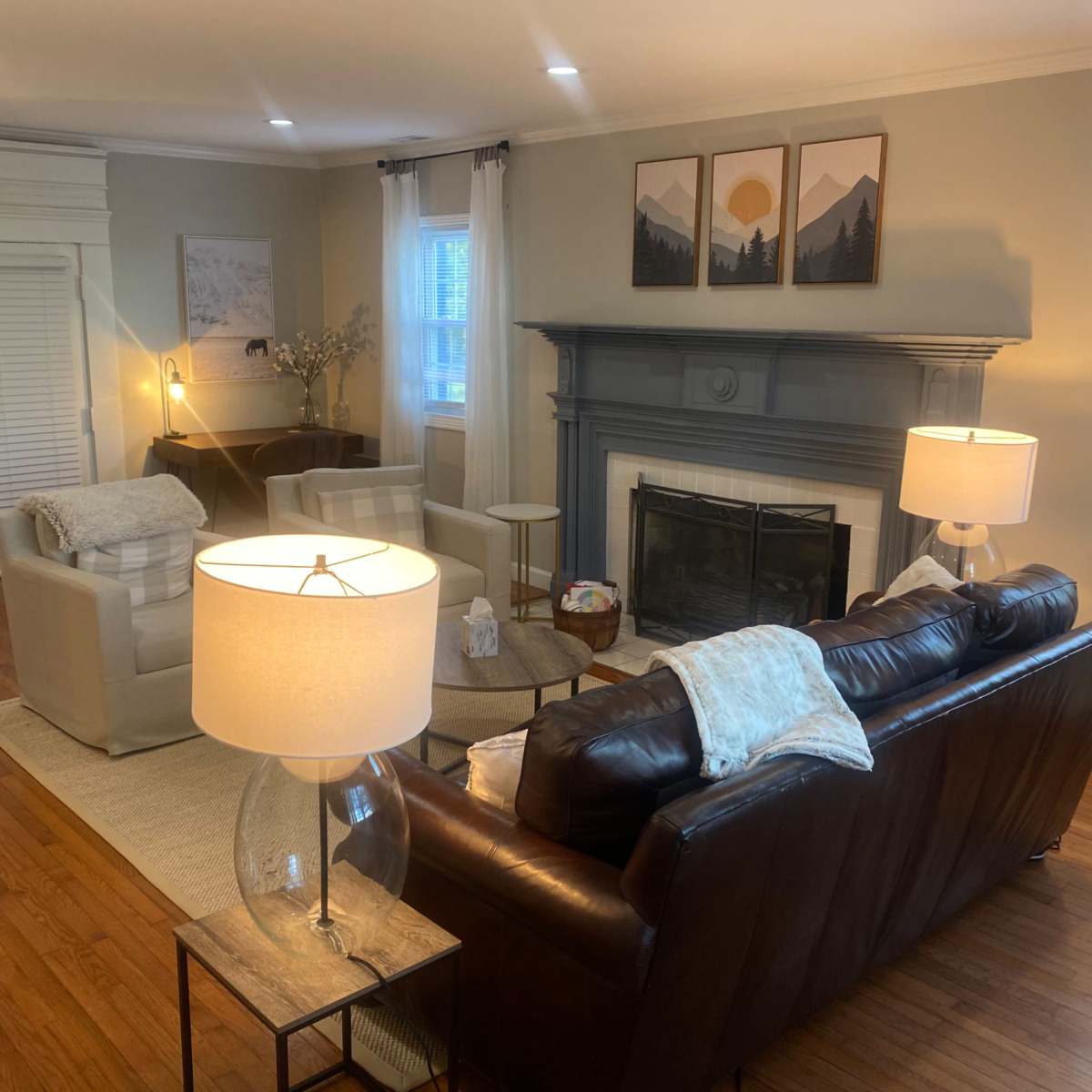 A living room features a brown leather sofa, two light-colored armchairs, a fireplace with a gray mantel, and lamps providing soft lighting.