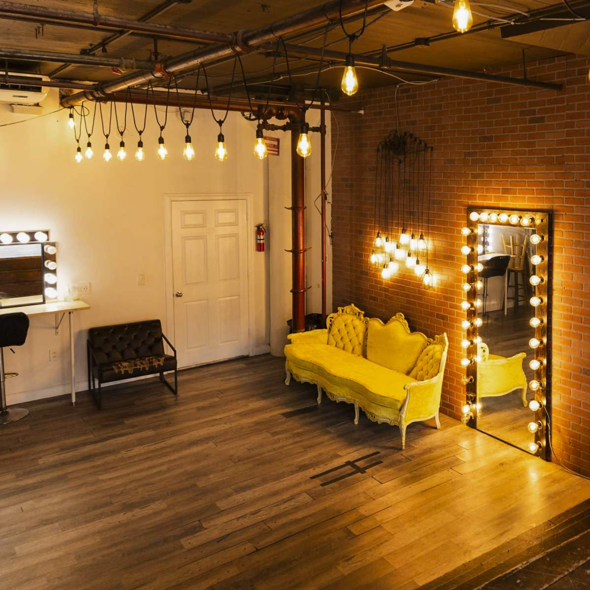 A stylish room with yellow sofas, a large mirror framed by lights, and exposed brick walls.