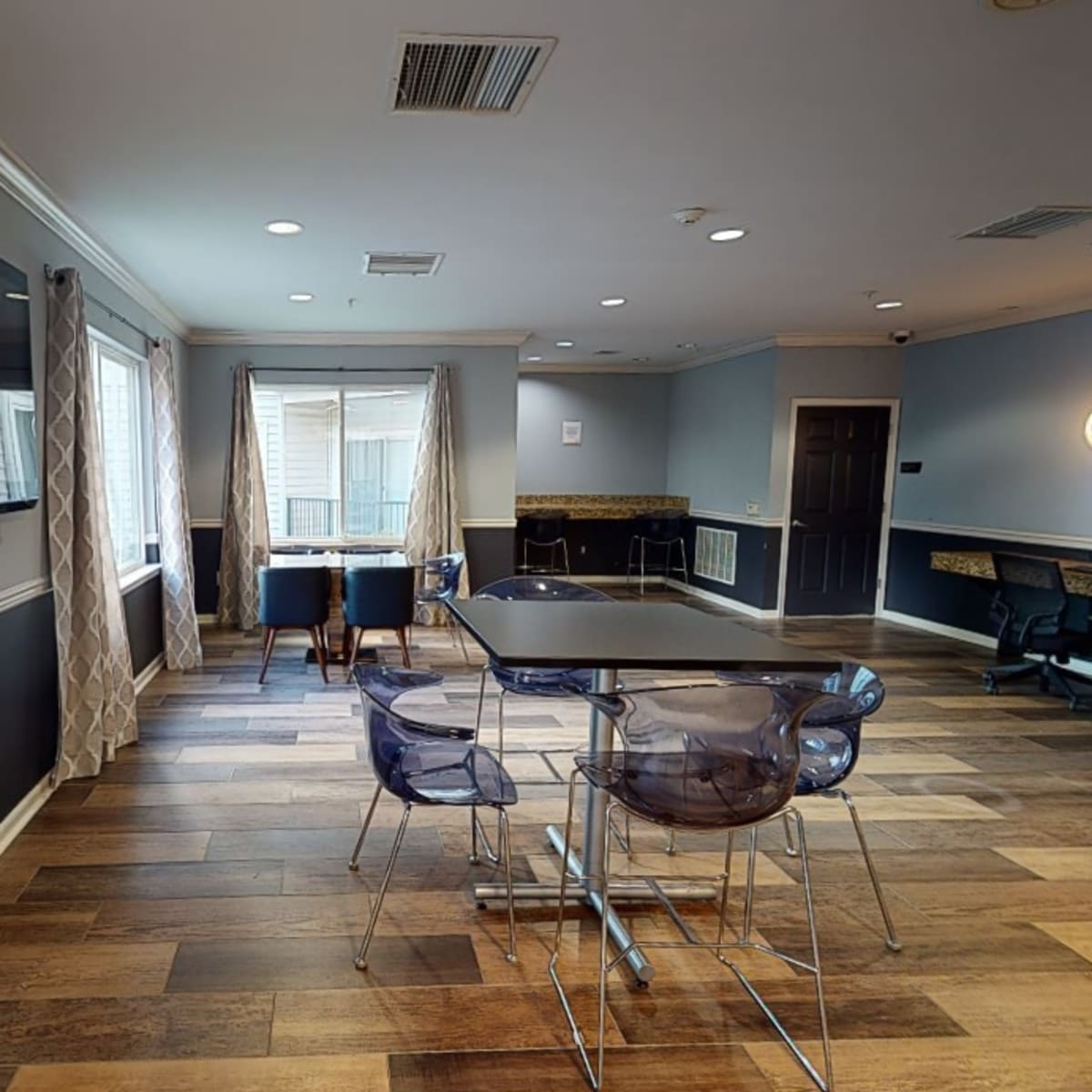 A modern room features a large table surrounded by transparent chairs, with a television on one wall and workstations in the background.