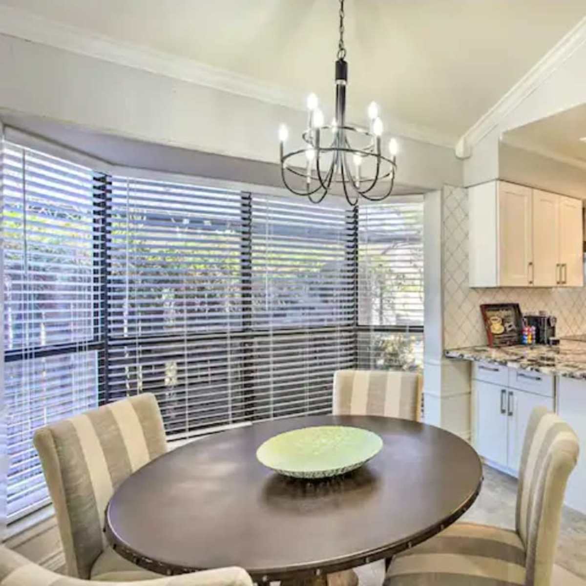 A modern kitchen and dining area with a round table, four chairs, and large windows covered with blinds.
