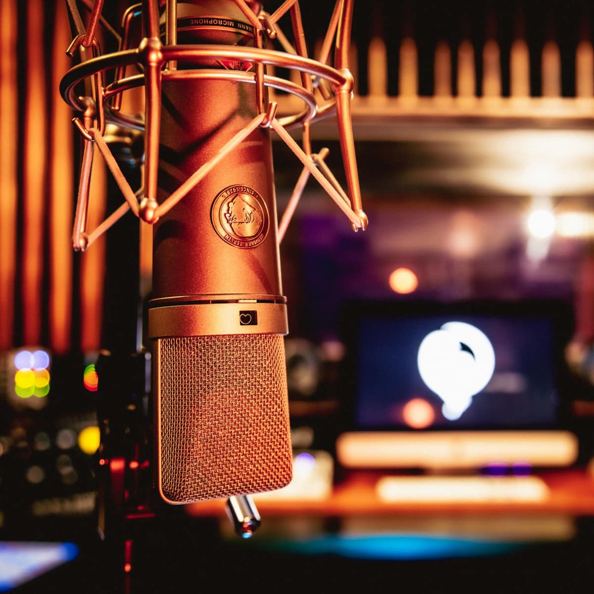 A microphone is positioned prominently in a sound studio with audio equipment and a computer in the background.