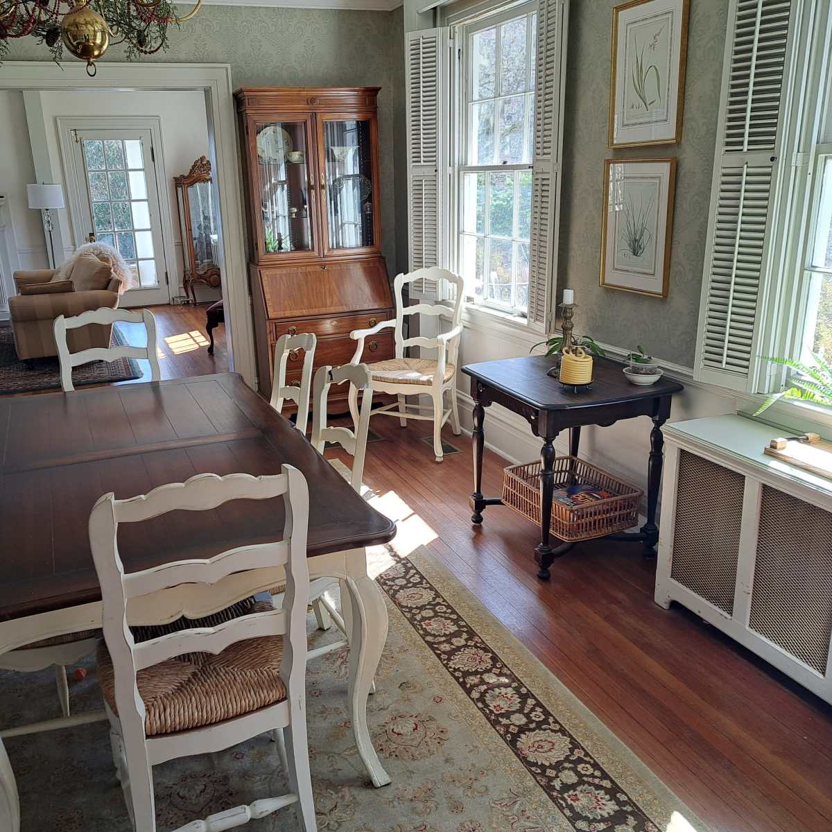 A sunlit dining room with a wooden table surrounded by white chairs, a sideboard, and large windows featuring decorative plants.