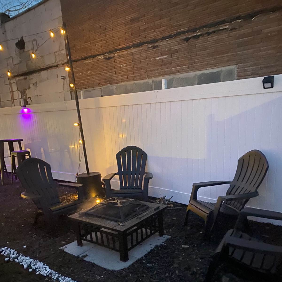 Four Adirondack chairs positioned around a fire pit in a dimly lit outdoor setting, with string lights illuminating the area.