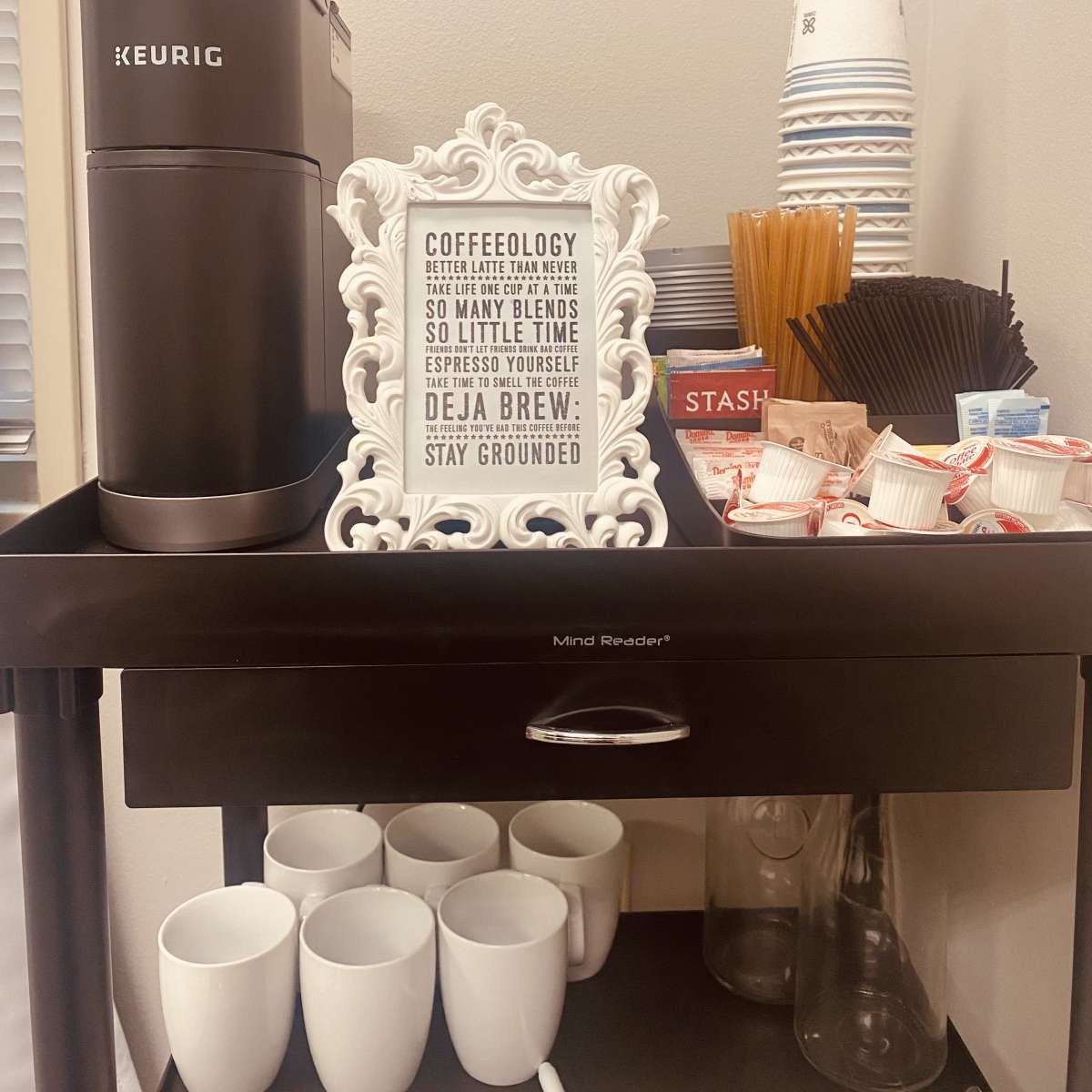 A coffee station with a Keurig machine, cups, and a decorative sign with coffee-related sayings.