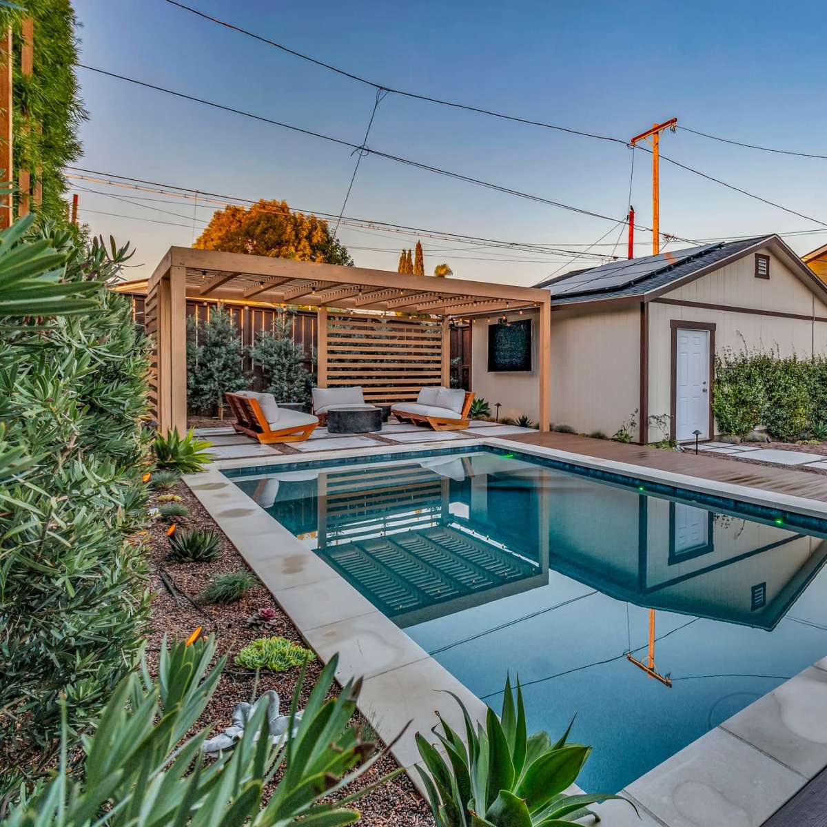 A rectangular swimming pool surrounded by wooden decking, lounge chairs, and landscaped greenery, with a shed and house in the background.