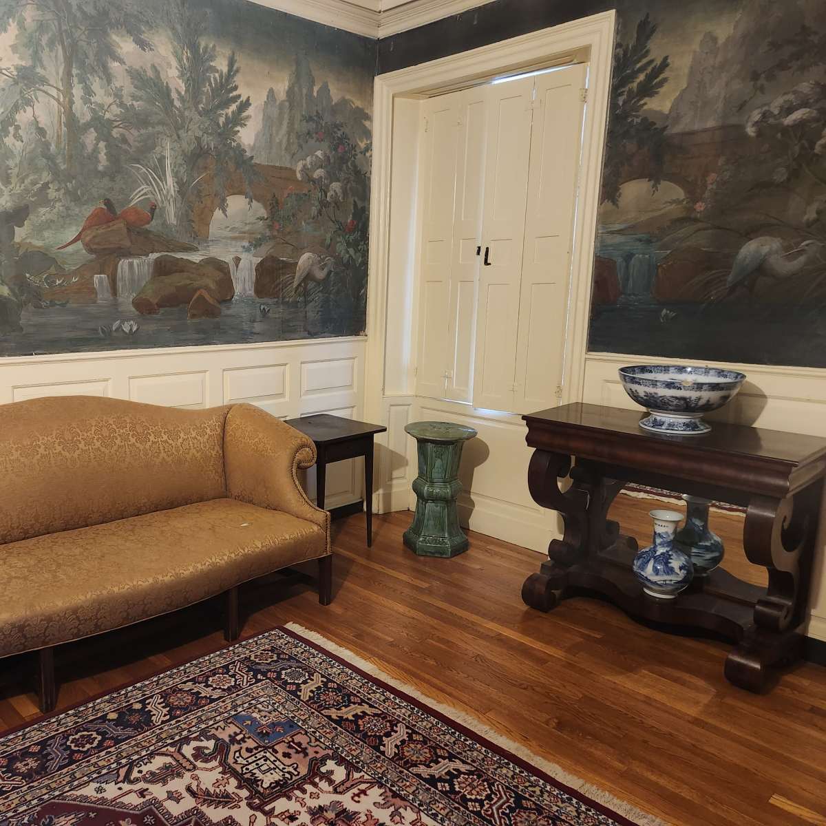 A room with a patterned couch, a wooden table, and an ornately painted mural featuring birds and a natural scene on the walls.