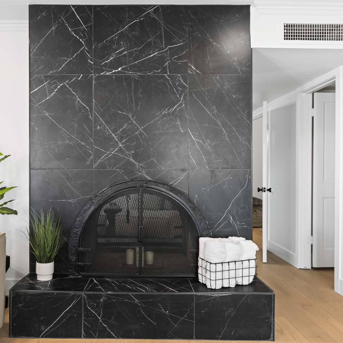 A black marble fireplace with a curved iron grate, surrounded by a light-colored living space featuring a plant and a basket of towels.
