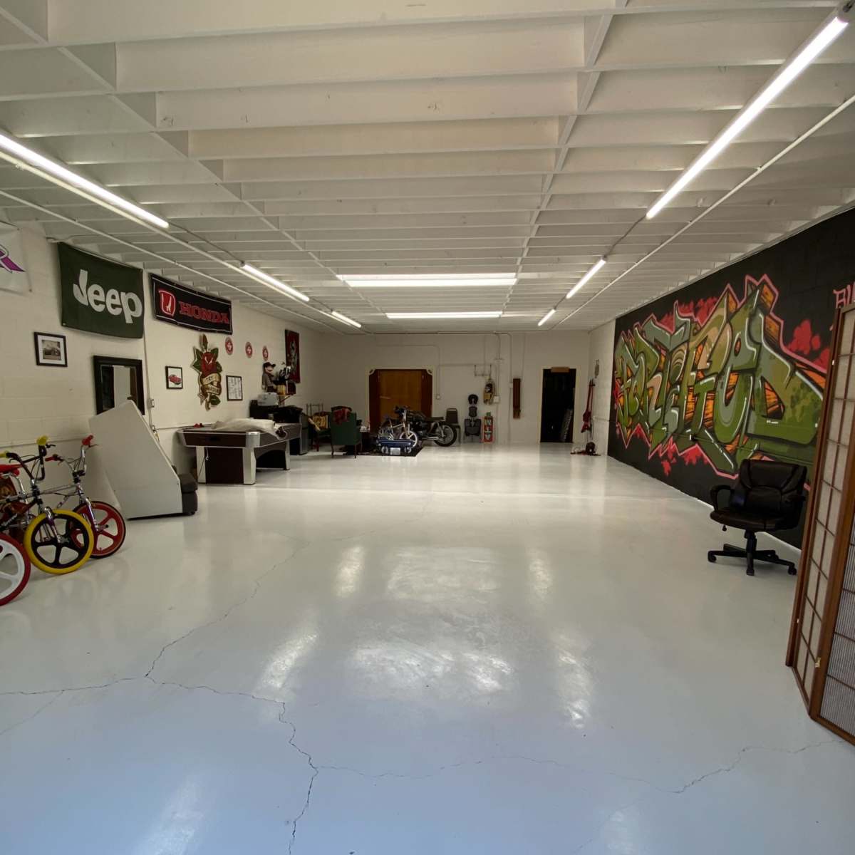 A spacious garage or workshop with a polished white floor, featuring bikes, motorcycles, and graffiti art on the walls.