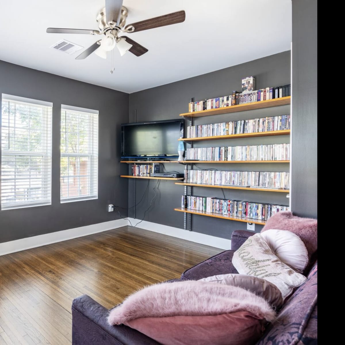 A small room with a window, a ceiling fan, a purple couch, bookshelves filled with DVDs, and a wooden chair near a desk.