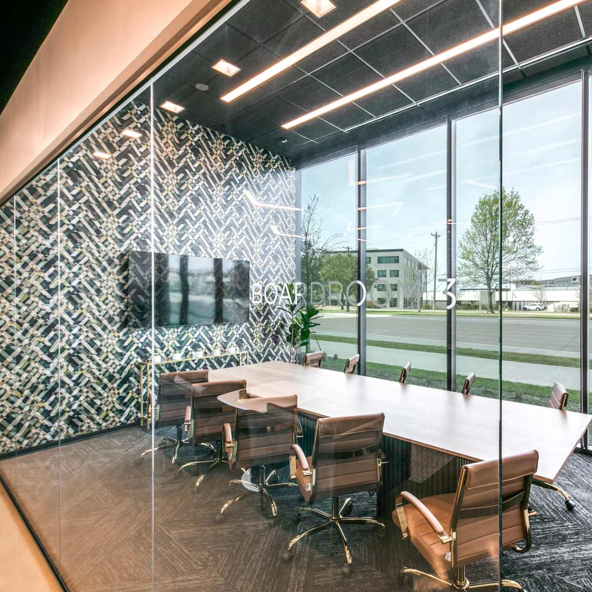 A modern conference room features a large table surrounded by chairs, with a patterned wall and large windows overlooking an outdoor area.