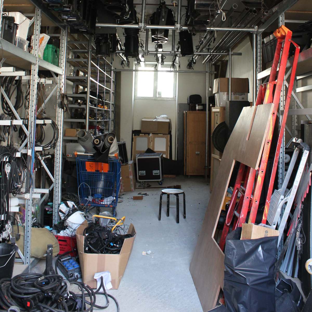 A cluttered storage room filled with lighting equipment, tools, and various materials organized on metal shelving and scattered on the floor.