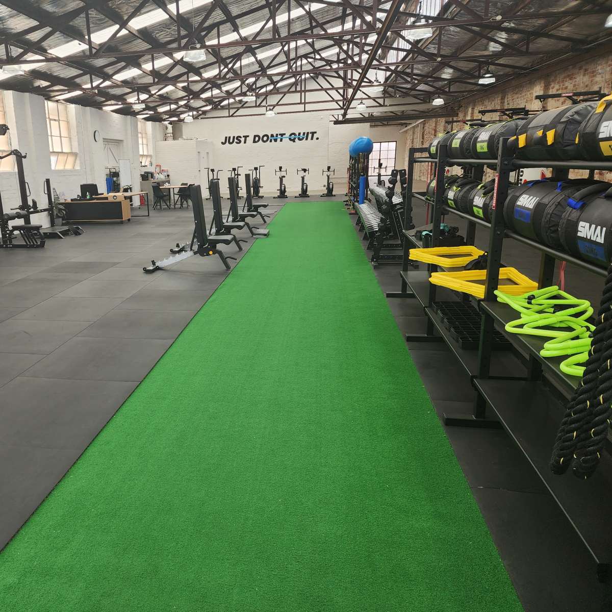 A spacious gym with a green artificial turf area, various fitness equipment, and a motivational sign on the wall that reads "JUST DON'T QUIT."