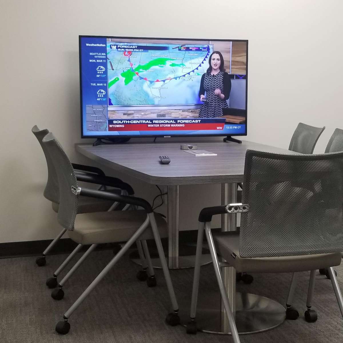 A conference room features a large television displaying a weather forecast, with several chairs arranged around a table.