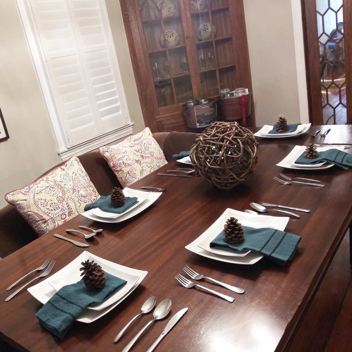 A dining table is set with plates, utensils, and decorative pinecones, surrounded by cushioned chairs and a cabinet with glassware in the background.