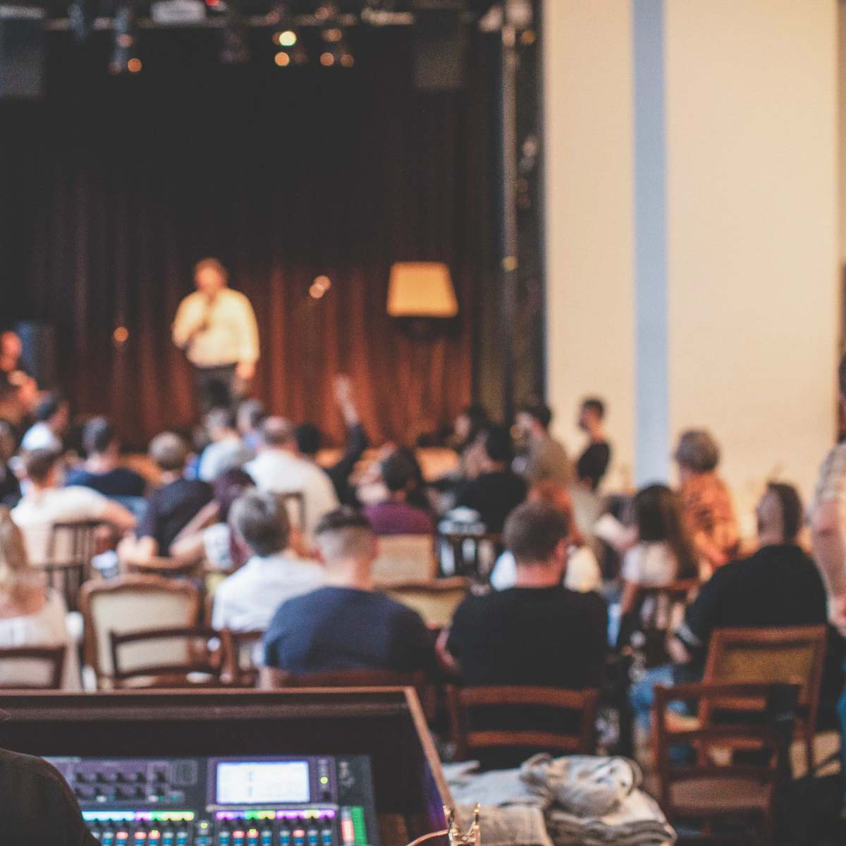 A crowd of people sits in a dimly lit venue, watching a performer on stage while a sound technician adjusts equipment in the foreground.