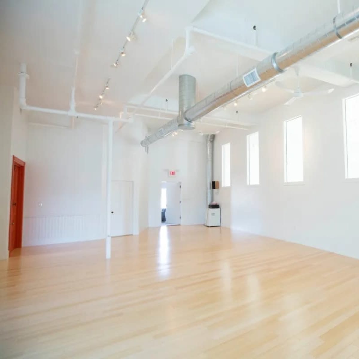 A spacious, empty room with wooden flooring, white walls, and large windows allowing natural light.