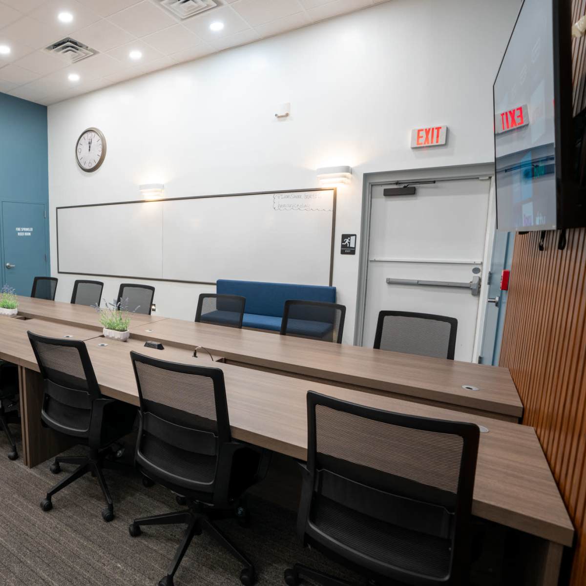 A conference room features a long, rectangular table with black chairs, a whiteboard, a clock on the wall, and a doorway leading to a restroom.