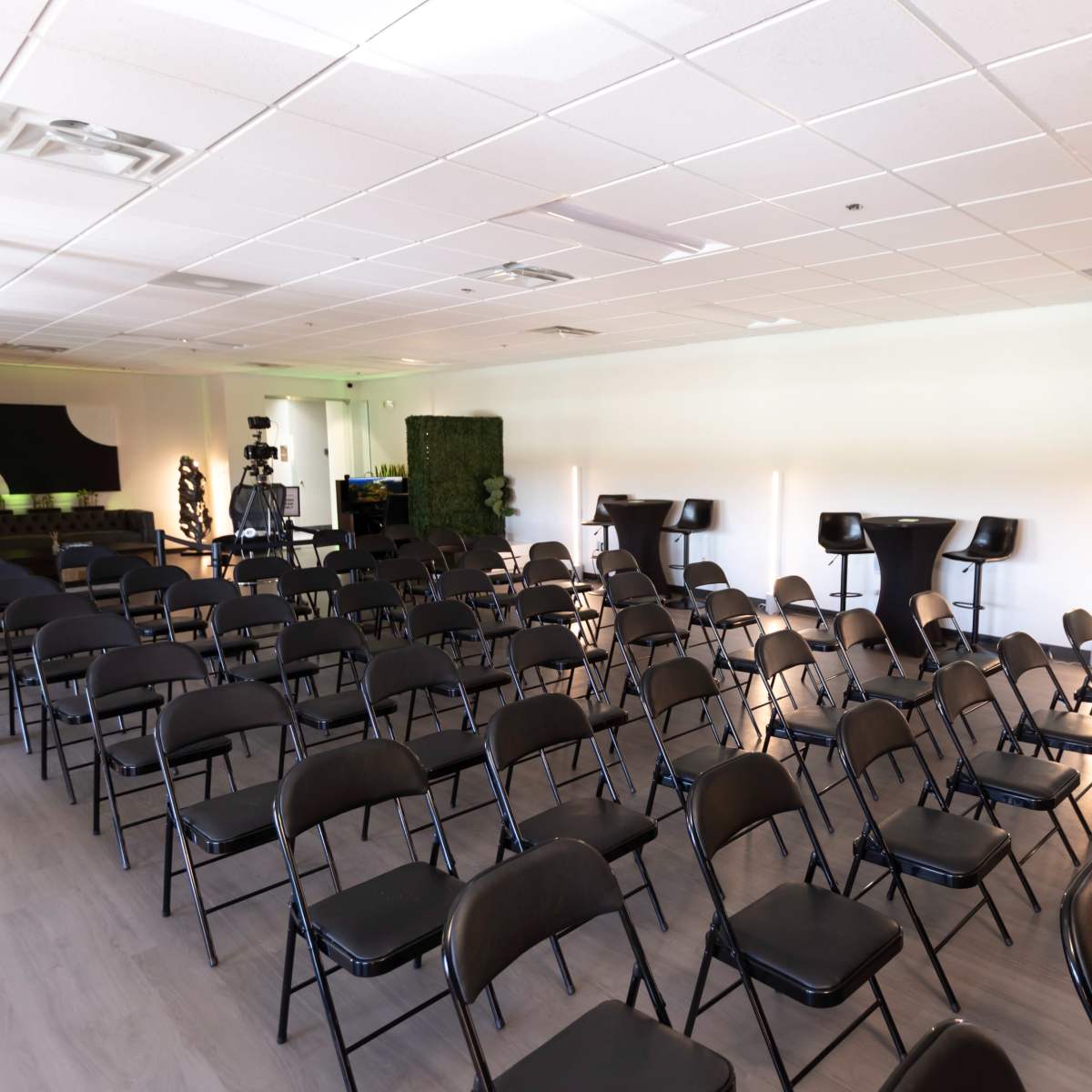 A neatly arranged room with rows of black folding chairs facing a presentation area and a few high-top tables along the sides.