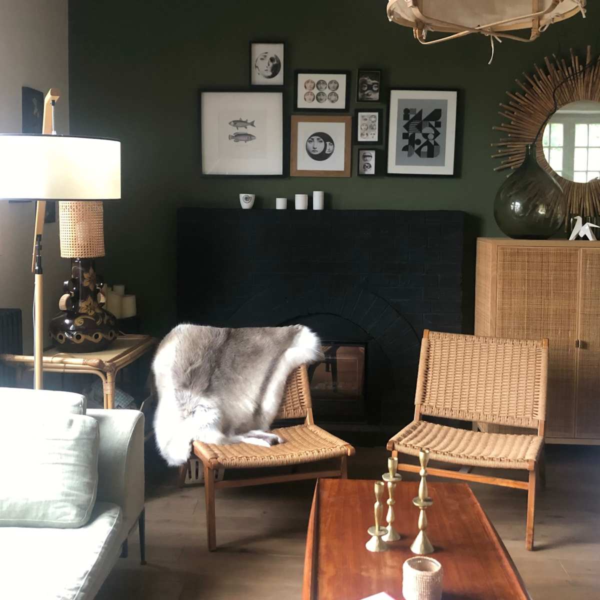 A cozy living room with a dark green wall, a black fireplace, wicker chairs, a wooden coffee table, and various framed artworks on display.